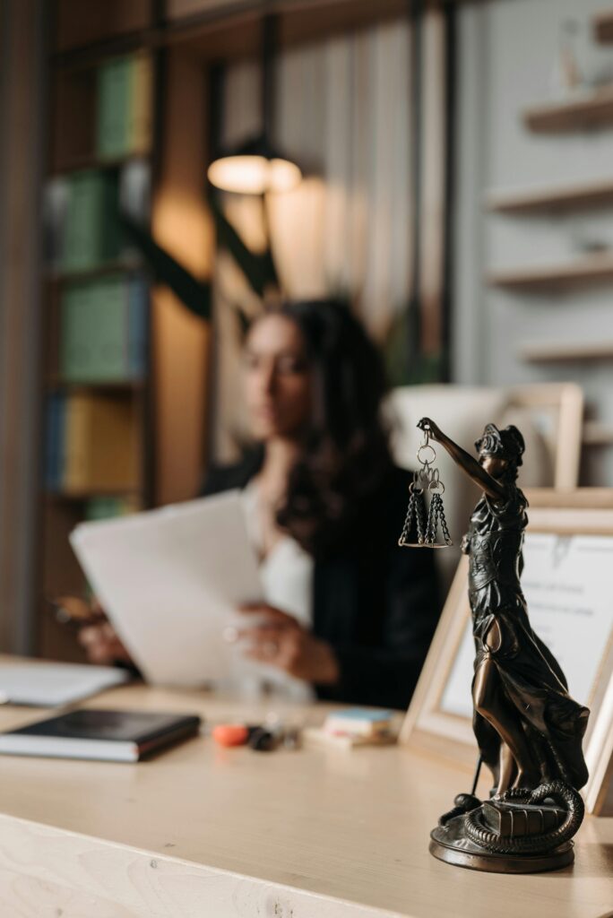pexels-photo-8111815-8111815 Close-up of a justice figurine on desk with blurred female lawyer in office setting.